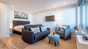 A modern living room with a grey sofa, a matching armchair, a small ottoman, a flat-screen TV, and a painting on the wall.