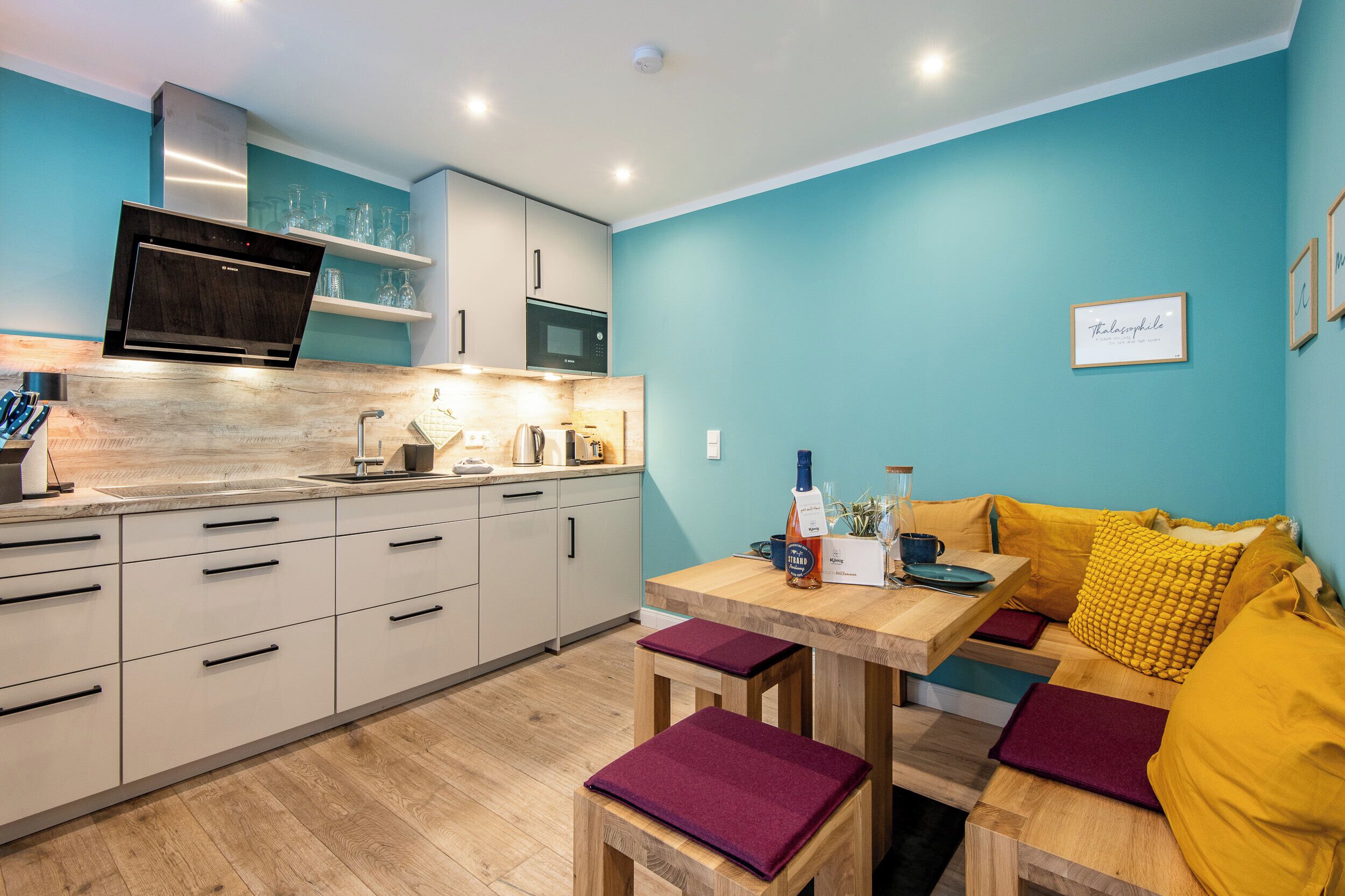 A modern kitchen with white cabinets, a stainless steel range hood, and a microwave. There is a wooden table with a bench and yellow cushions.