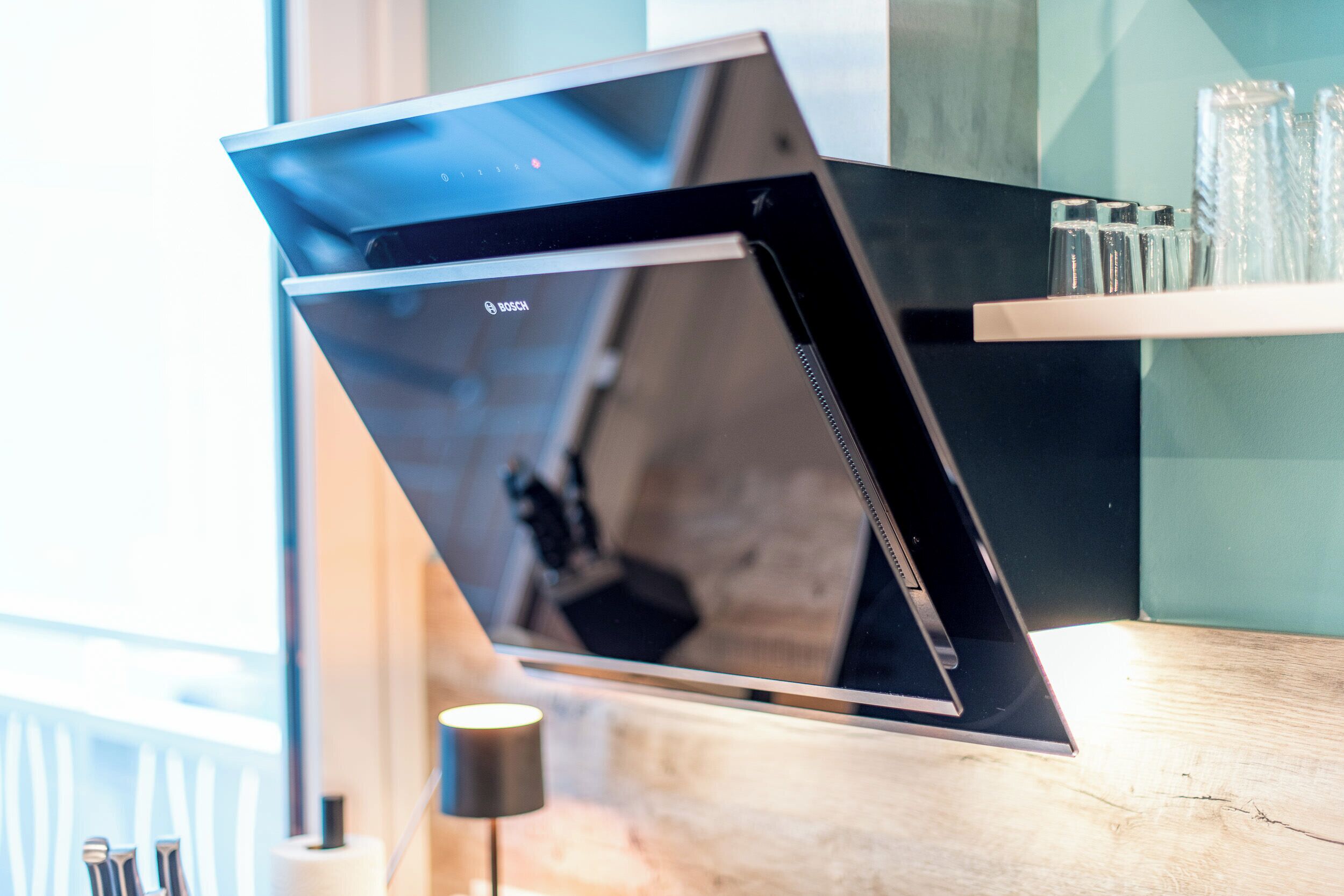 A modern kitchen with a Bosch range hood, a wooden countertop, and a glass shelf with glasses.