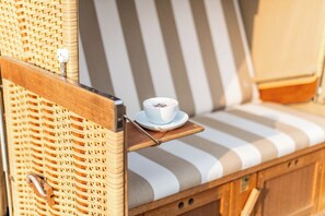 A wicker lounge chair with a striped cushion, a small table with a cup of coffee, and a power outlet on the backrest. - House Nordmarkgarten, Whg. 02a (Westerland)