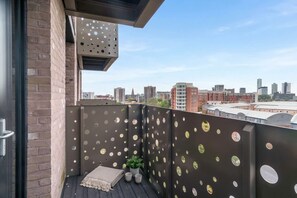 A balcony with a view of the city skyline, featuring a brick wall, a metal perforated panel, and a small potted plant. - Modern & Bright Central Liverpool 1 Bed Apartment (Liverpool)