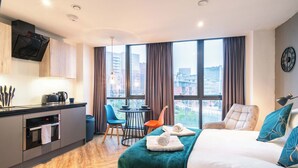 Moderni hotellihuone, jossa on pieni keittiö, turkoosinsinisellä tekstiilillä varustettu sänky, näköala kaupunkiin ja ruokailutila tuoleineen.