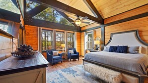 A spacious bedroom with a large bed, two armchairs, a wooden desk, and a large window offering a view of trees. - Games, Hot Tub, Fire Pit, Pets, Times Like These (Broken Bow)