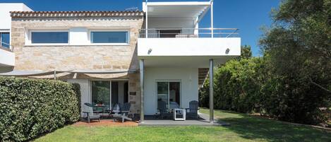 A modern two-story house with a patio area, outdoor seating, and a view of the surrounding greenery.
