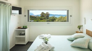A bedroom with a large window, a bed with rolled towels, a small shelf with books and a plant, and a view of a landscape. - Beach Apartments (Olbia)