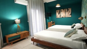 A bedroom with teal walls, a wooden bed frame, two beds, a bedside table with a lamp, a TV on the wall, and a window with curtains. - Ross Family Suites  (Reggio Calabria)