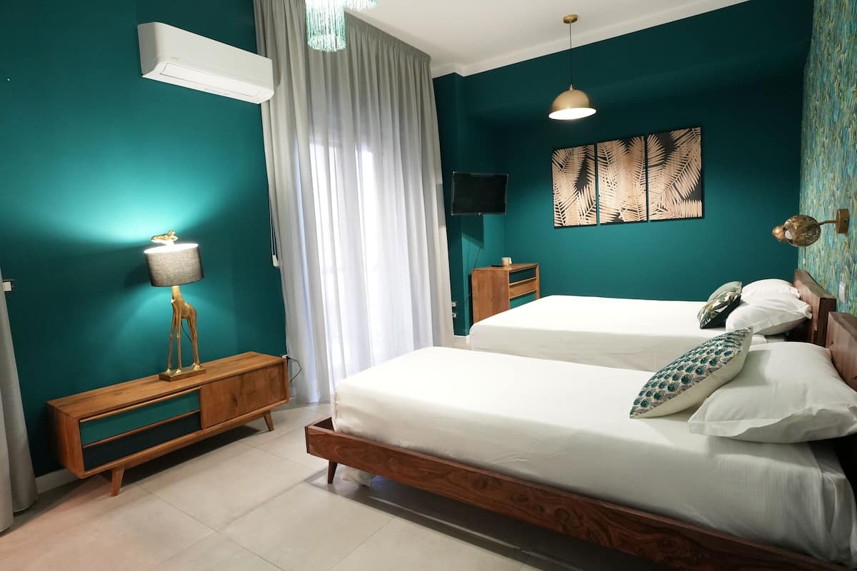 A bedroom with teal walls, a wooden bed frame, two beds, a bedside table with a lamp, a TV on the wall, and a window with curtains.