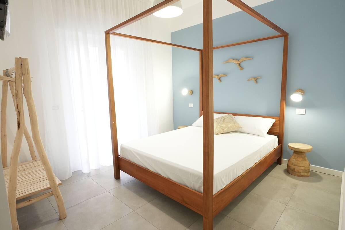 A four-poster bed with a wooden frame and white bedding, a wooden stool, and wall-mounted lights in a room with light blue walls and a white curtain.