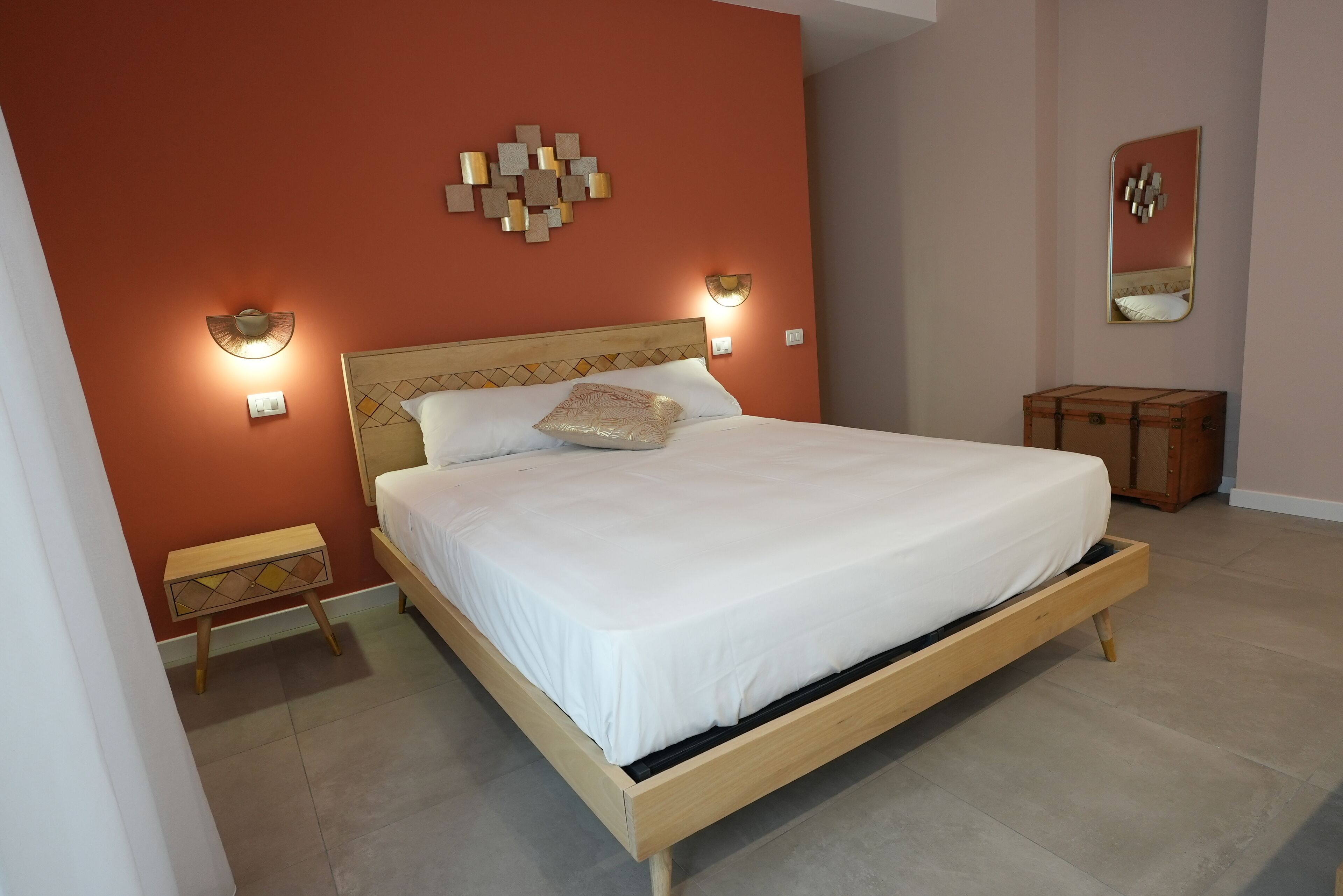 Un dormitorio con cama de madera, dos mesitas de noche, una mesita auxiliar, un espejo montado en la pared y apliques decorativos.