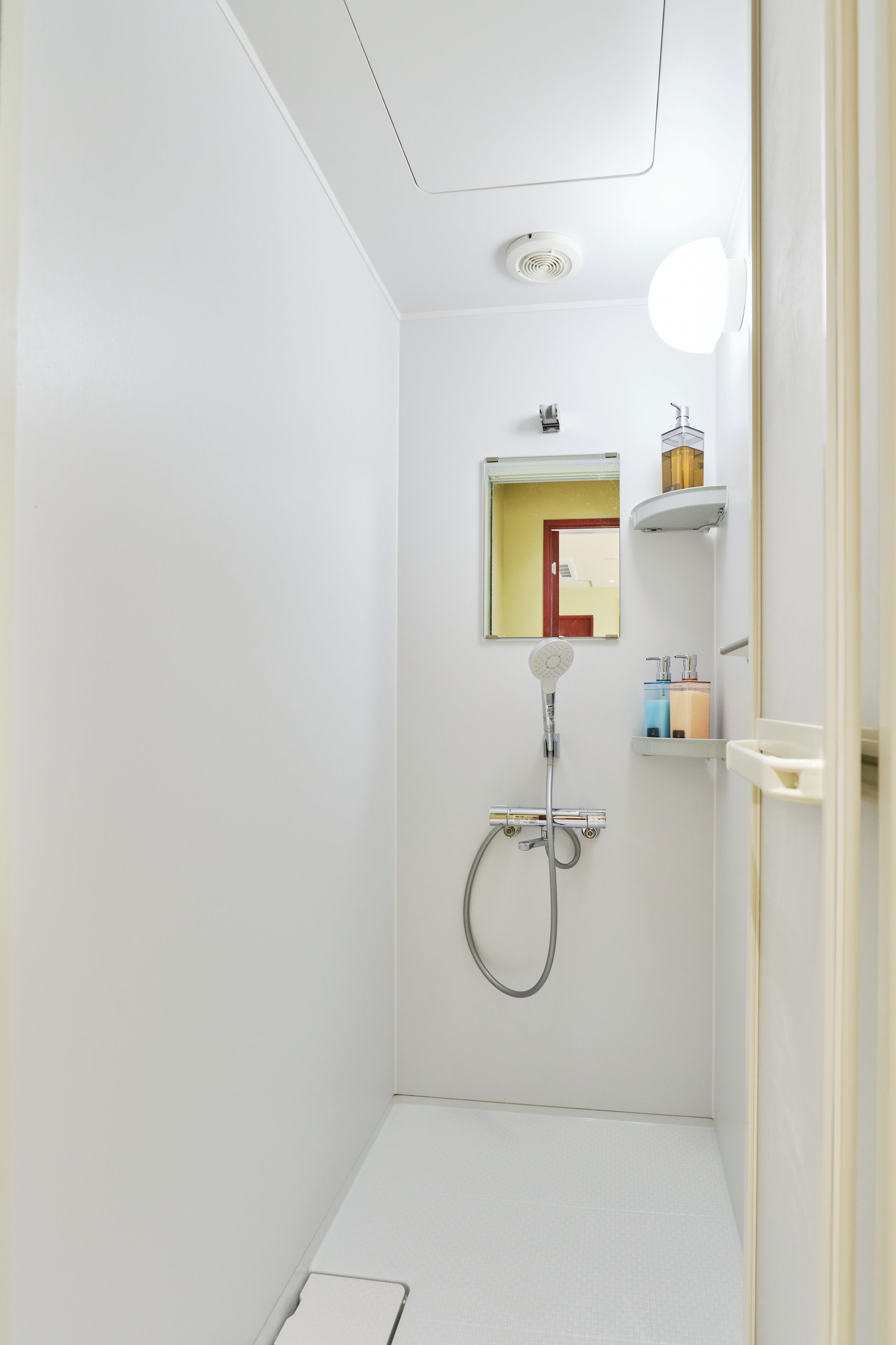 A small, white bathroom with a shower, a mirror, and a shelf with toiletries.