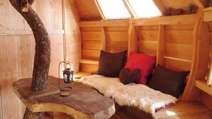 A cozy cabin with a sloped roof, wooden walls, a fur-covered sofa, and a tree trunk table with a lantern. - Cabane perchée Arbràmâne (5 people) (Vittel)