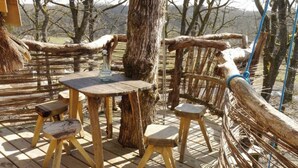 A rustic wooden deck with a round table and four stools, surrounded by natural tree trunks and branches. - Cabane perchée Arbràmâne (5 people) (Vittel)