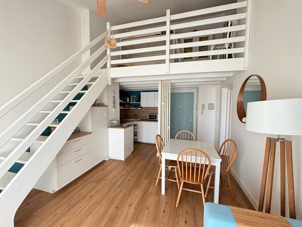 A loft-style room with a staircase, a dining area with wooden chairs, a kitchen with white cabinets, and a blue door.