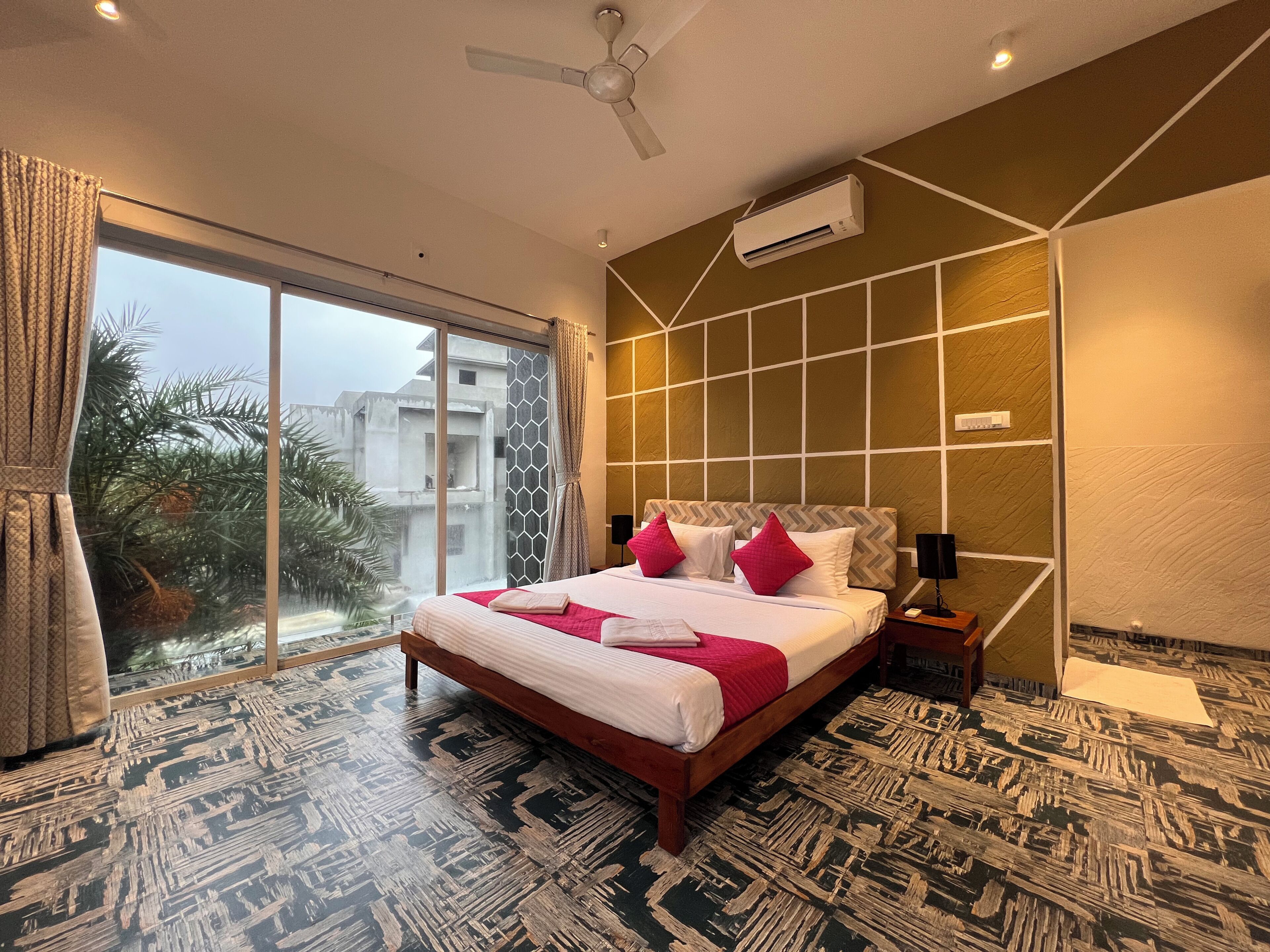 A hotel room with a large bed, a ceiling fan, an air conditioning unit, a nightstand, and a view of buildings outside.