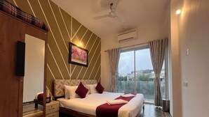 Hotelkamer met een bed, nachtkastje, spiegel, plafondventilator, airconditioning en een groot raam met gordijnen.