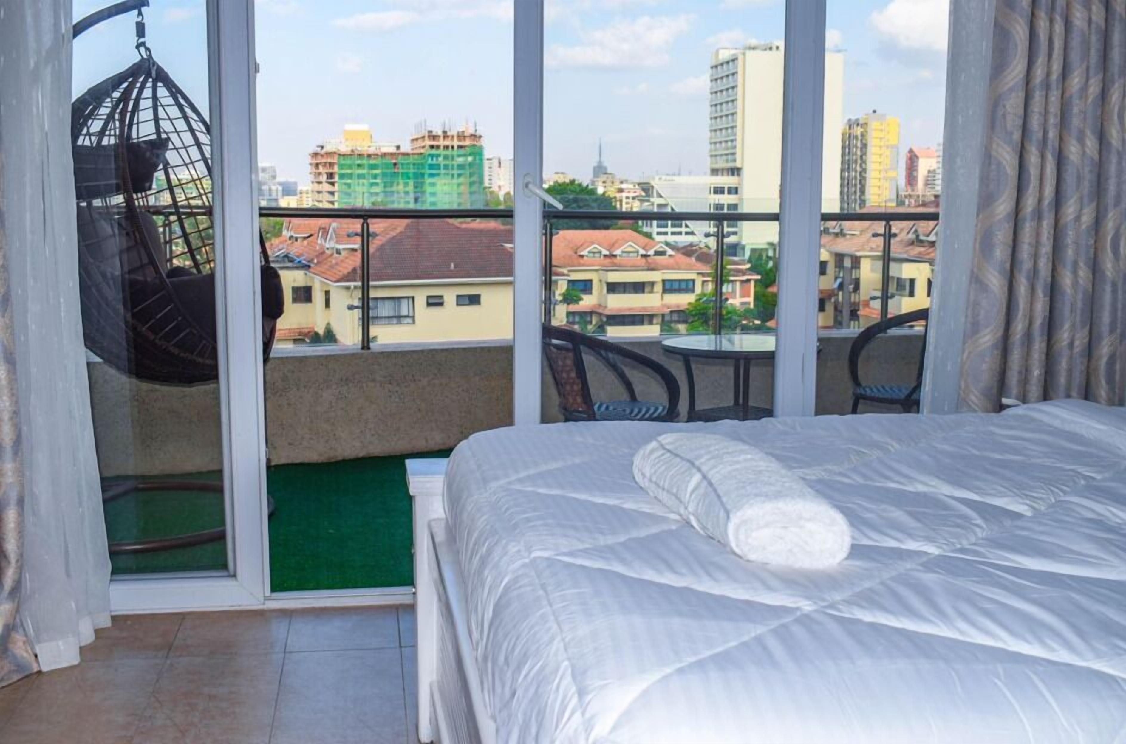Un balcone con una sedia a dondolo, un letto con copripiumino bianco e vista sugli edifici della città.