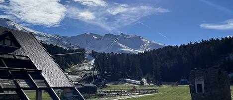 Un chalet de montagne avec un toit en pente, un petit bâtiment sur lequel on peut voir une enseigne, et une vue dégagée sur des montagnes enneigées baignées par un soleil éclatant.