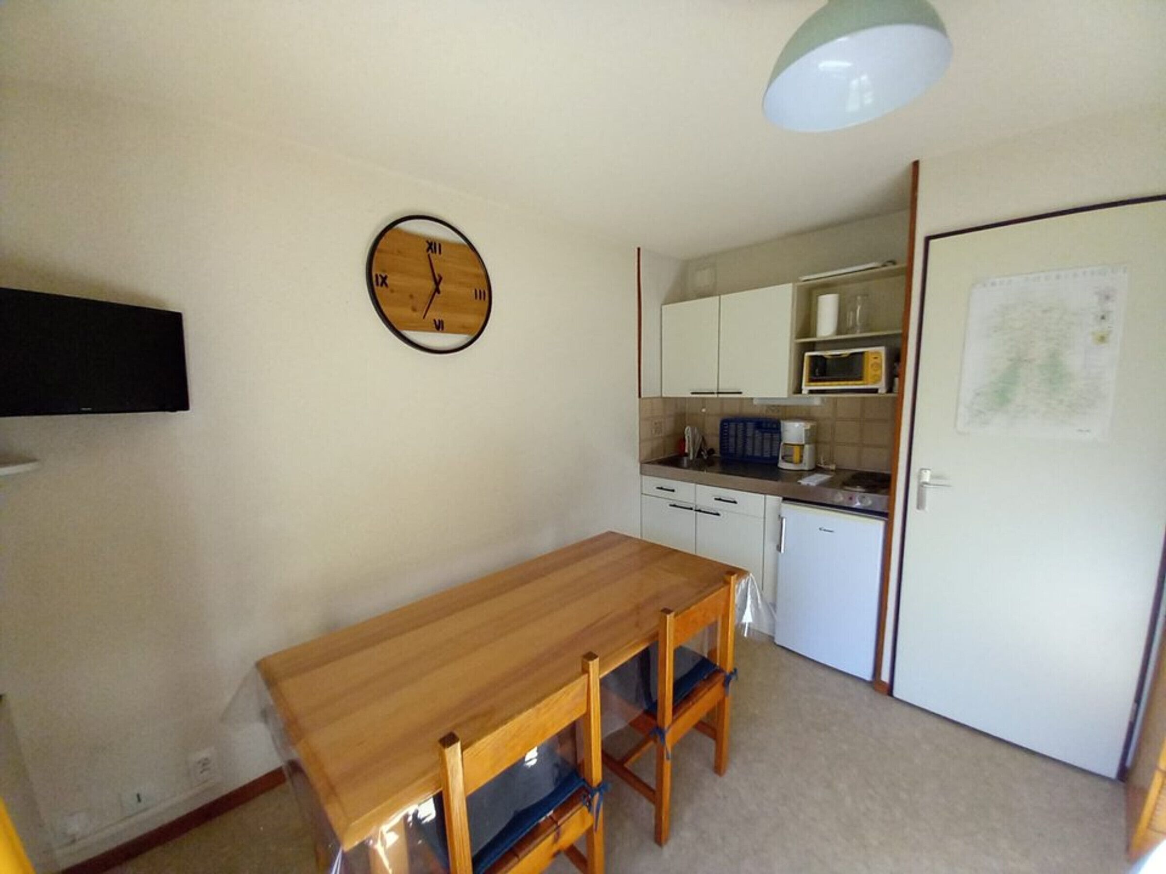 Una pequeña cocina con una mesa y sillas de madera, un reloj en la pared y un microondas.