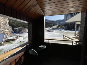 Une cabane en bois dotée d’un balcon donnant sur un paysage enneigé, avec une piste de ski et des bâtiments visibles au loin.