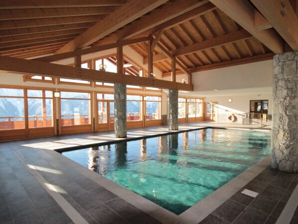 An indoor swimming pool with stone pillars, wooden beams, and large windows offering mountain views. - Spacious apartment, panoramic view (Villard-sur-Doron)