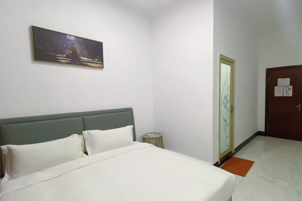 A hotel room with a bed, a painting on the wall, a round side table, and a door with a glass panel.