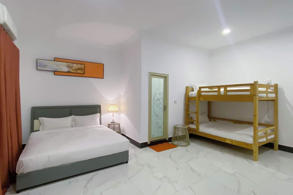 A hotel room with a single bed, a bunk bed, a bedside lamp, a framed artwork, and a door with a glass panel.