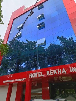 A modern building with a reflective glass facade, a prominent red and white sign indicating a restaurant and banquet facility, and a visible entrance with a parking sign. - Hotel Rekha International (Rajgir)