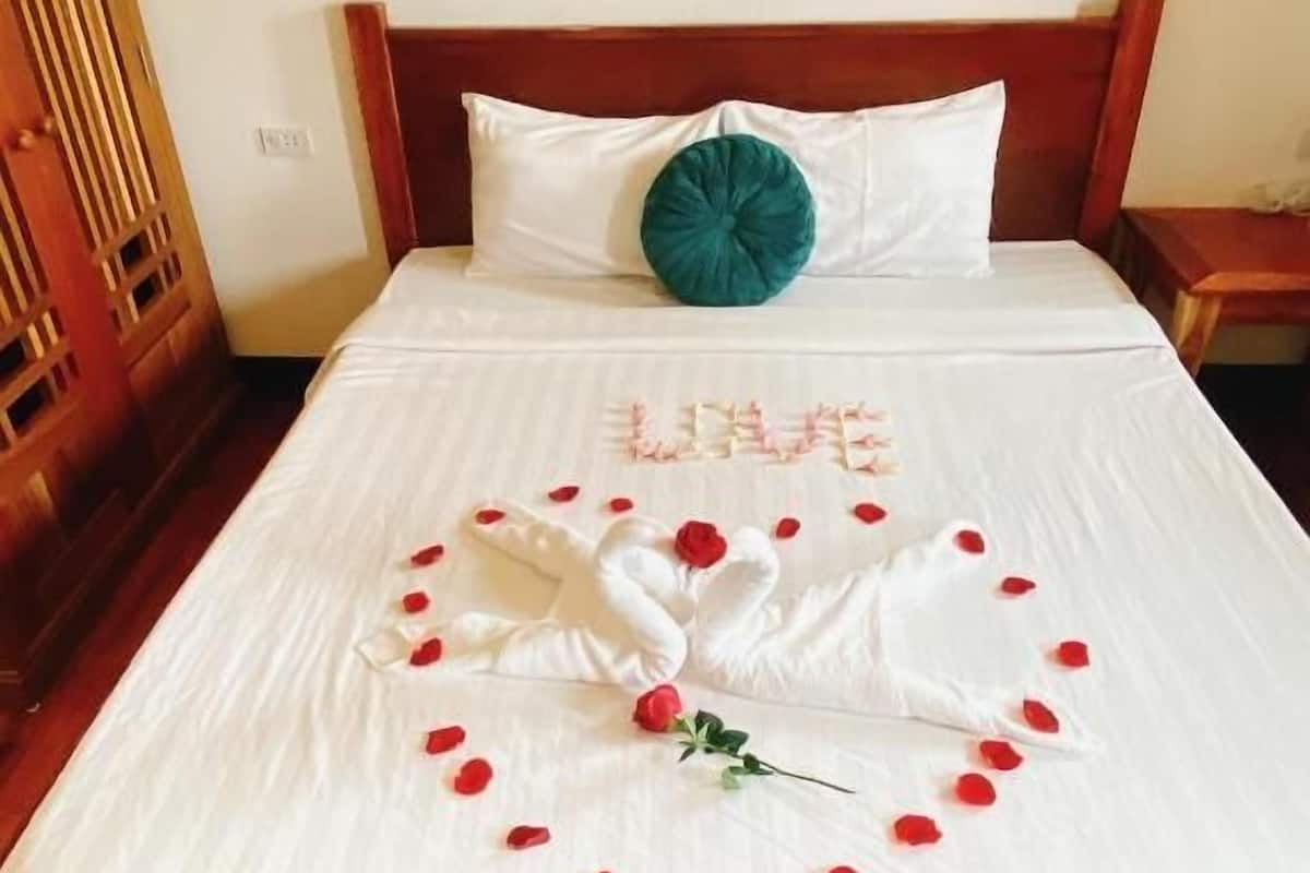 A neatly made bed with a heart-shaped arrangement of rose petals and a swan design, a teal pillow, and a white pillow on a wooden bed frame.
