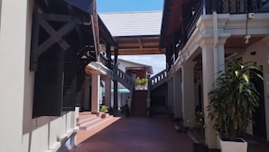 A covered walkway with a tiled floor, flanked by buildings with balconies and columns, leading to a covered area with a view of the sky.