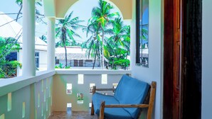 A balcony with a blue cushioned chair, a tiled floor, and a view of palm trees and buildings. - Saba house 24 (Paje)