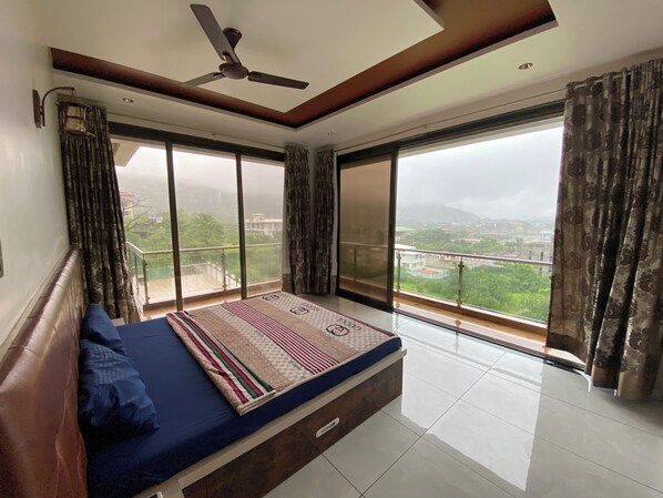 Un dormitorio con cama, ventilador de techo, ventanas amplias y vista a un paisaje urbano.