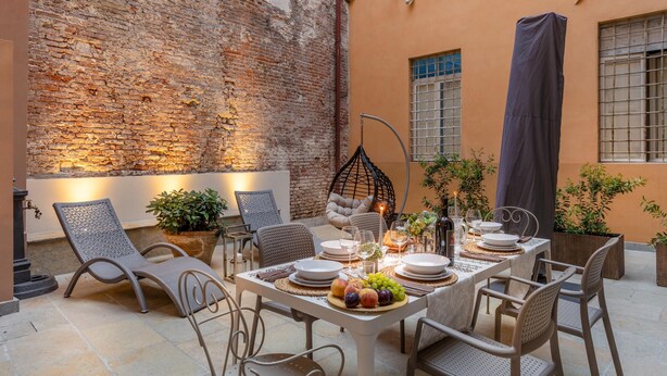 An outdoor dining area with a brick wall backdrop, wicker chairs, a table set with plates, glasses, and a fruit centerpiece. - Cortile Moderno Apartment (Lucca)