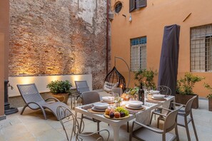 Un espacio de comedor al aire libre con un fondo de pared de ladrillo, sillas de mimbre, una mesa dispuesta con platos, vasos y un centro de mesa con frutas.