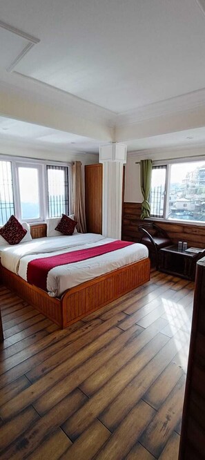 A hotel room with a large bed, wooden flooring, a small table, and a view of the city through large windows. - Hotel Apsara By GRB (Shimla)
