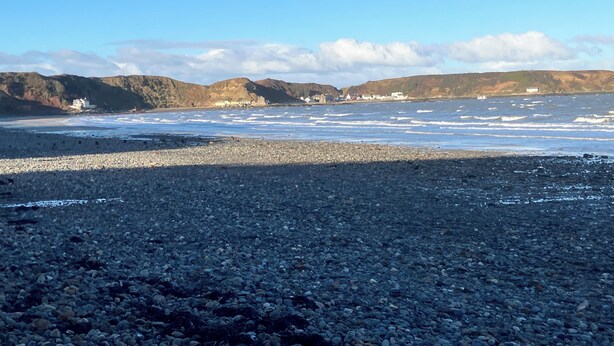 Une plage rocheuse où les vagues s’écrasent sur le rivage, une vue lointaine sur des collines et un ciel dégagé.