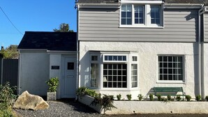 Une maison à deux étages, avec un toit gris, des murs extérieurs blancs et une porte d’entrée. On peut voir un banc et de petites plantes devant la maison.