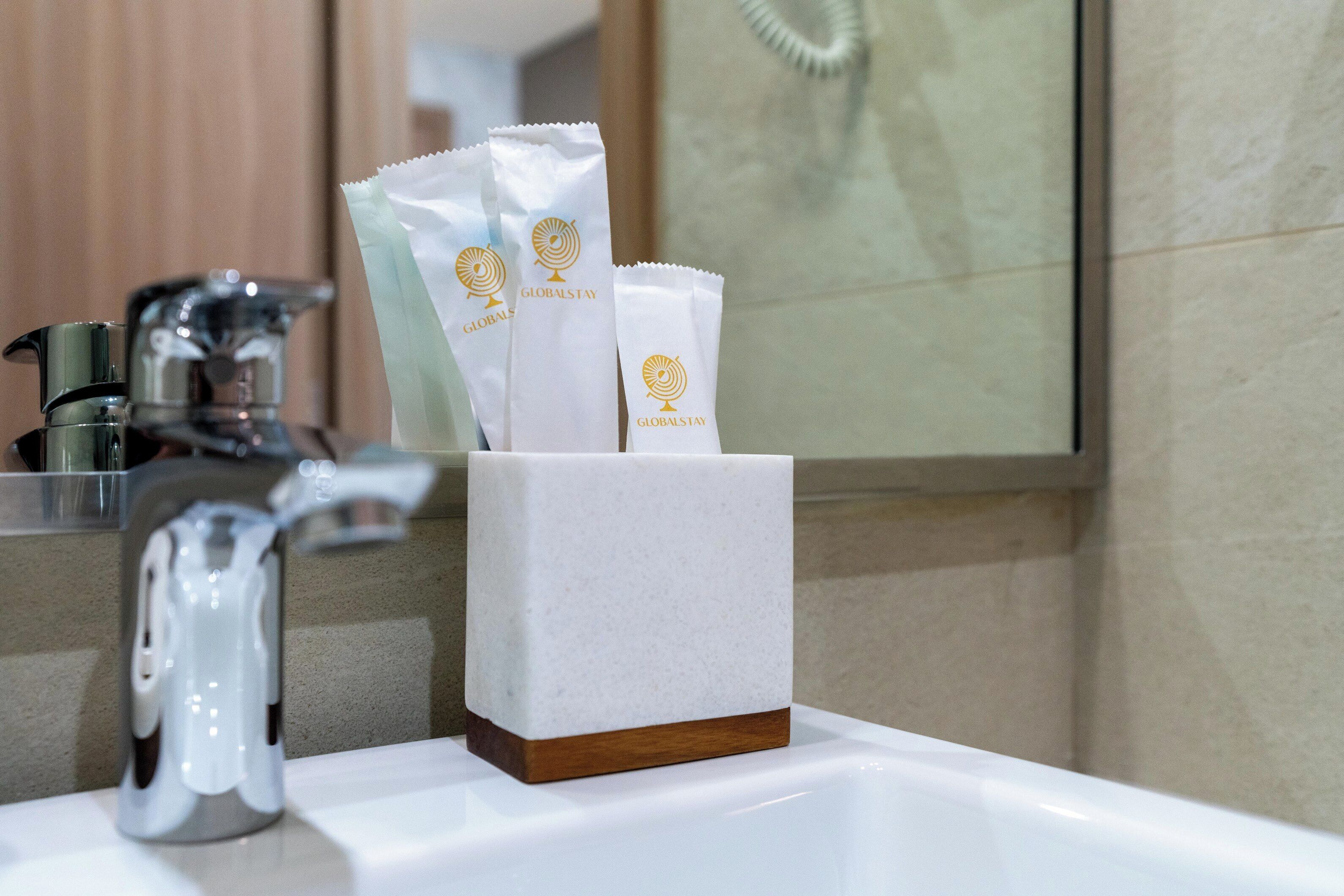 A bathroom sink with a white soap dispenser and a hand towel holder containing two white towels with a logo.