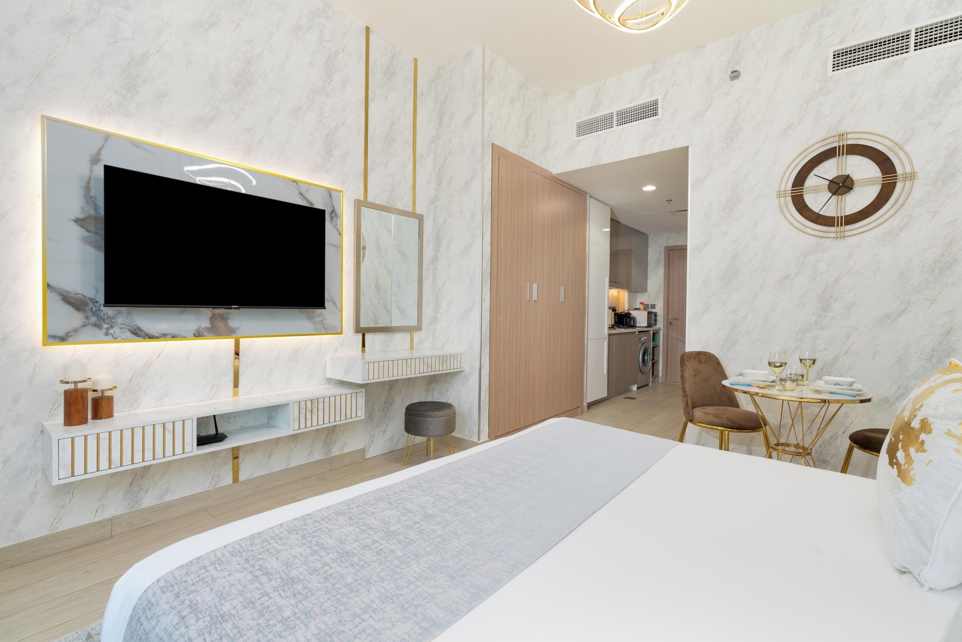 A modern hotel room with a large bed, a TV mounted on a marble wall, a small dining area with a table and chairs, and a separate kitchen area.