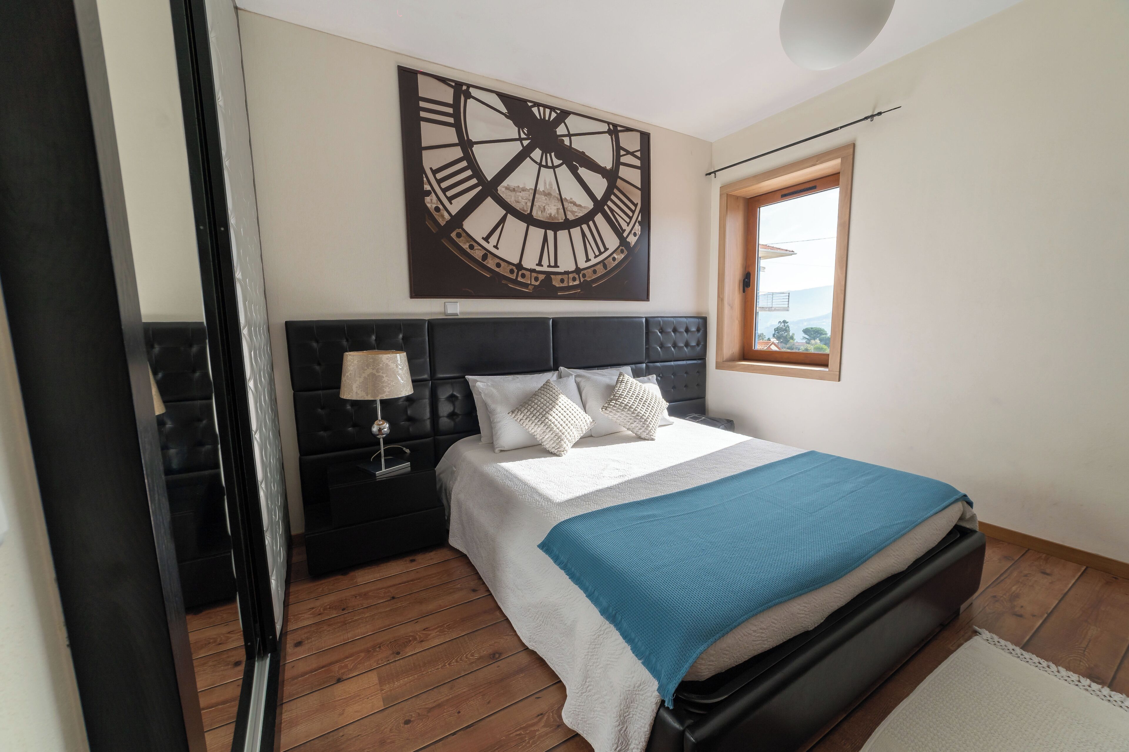 Ein Schlafzimmer mit einer großen Wanduhr, einem Bett mit blauer Decke, einem Nachttisch mit Lampe und einem Fenster mit Blick.