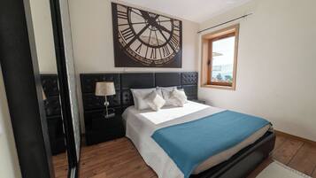 Ein Schlafzimmer mit einer großen Wanduhr, einem Bett mit blauer Decke, einem Nachttisch mit Lampe und einem Fenster mit Blick.