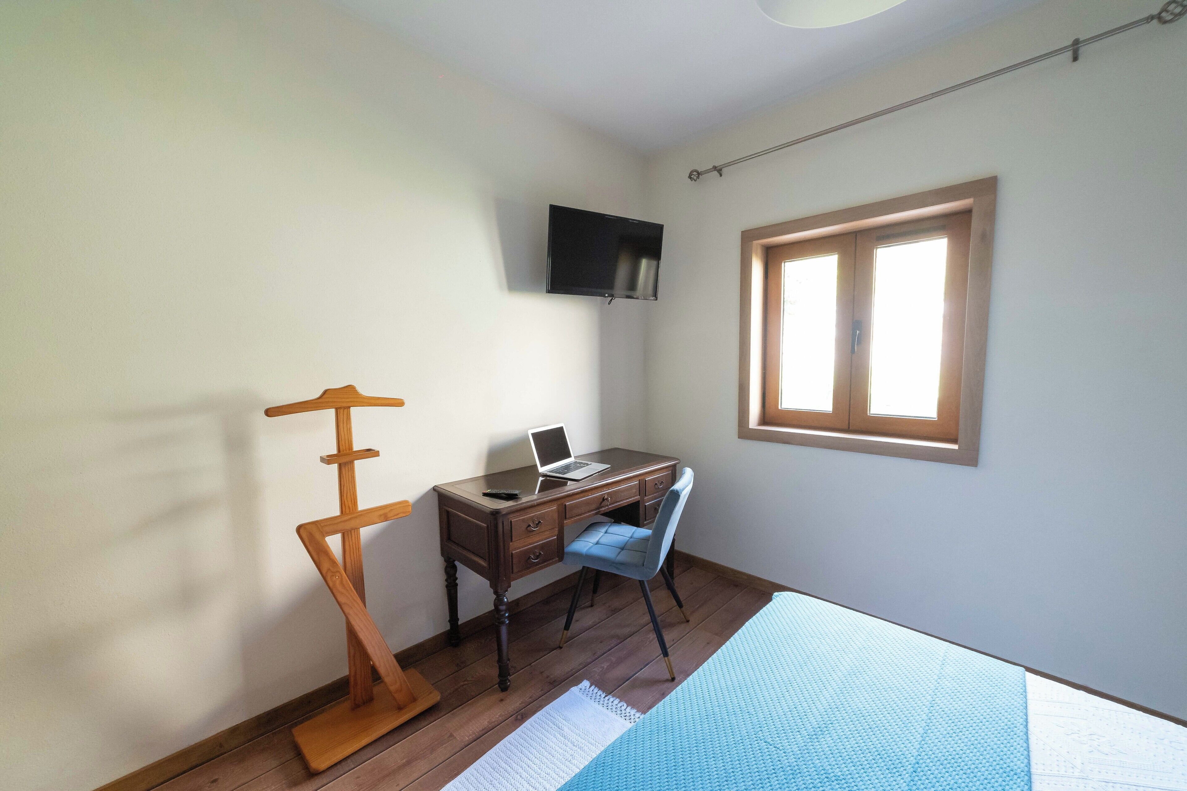 Een slaapkamer met een houten bureau, een laptop, een aan de muur bevestigde televisie en een raam met zonwering.