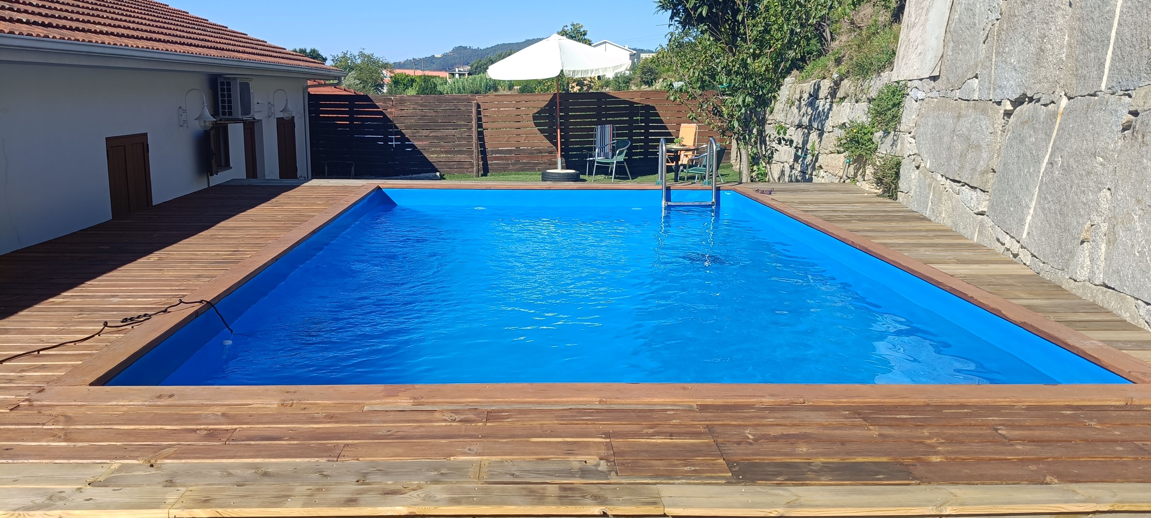Ein rechteckiger Swimmingpool mit umlaufender Holzterrasse, einem Holzzaun und einem Gebäude mit einem Ziegeldach im Hintergrund.