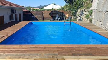 Ein rechteckiger Swimmingpool mit umlaufender Holzterrasse, einem Holzzaun und einem Gebäude mit einem Ziegeldach im Hintergrund.