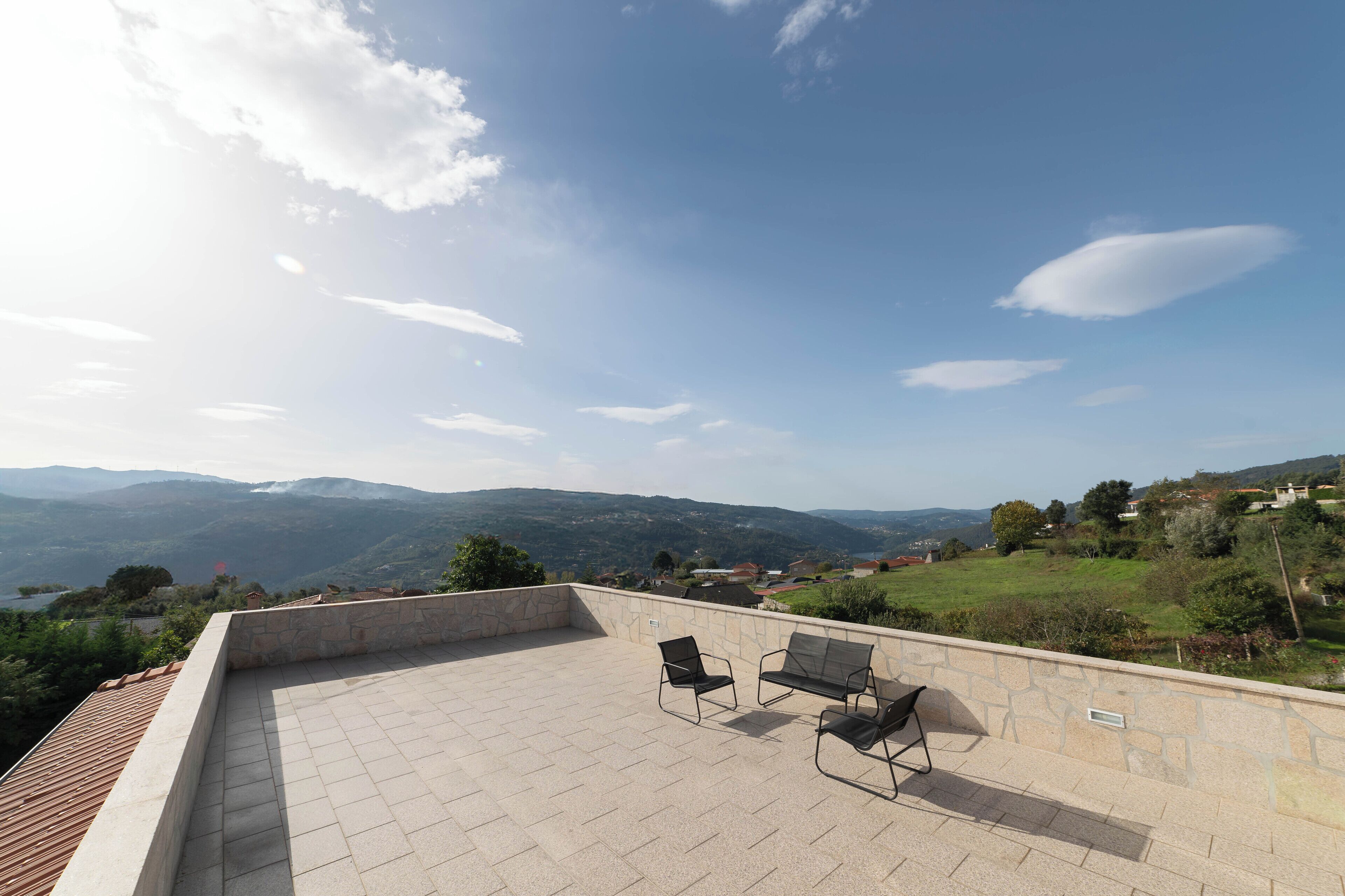 A rooftop terrace with stone walls and two black chairs, overlooking a valley with distant mountains and a body of water.