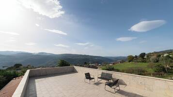 A rooftop terrace with stone walls and two black chairs, overlooking a valley with distant mountains and a body of water.