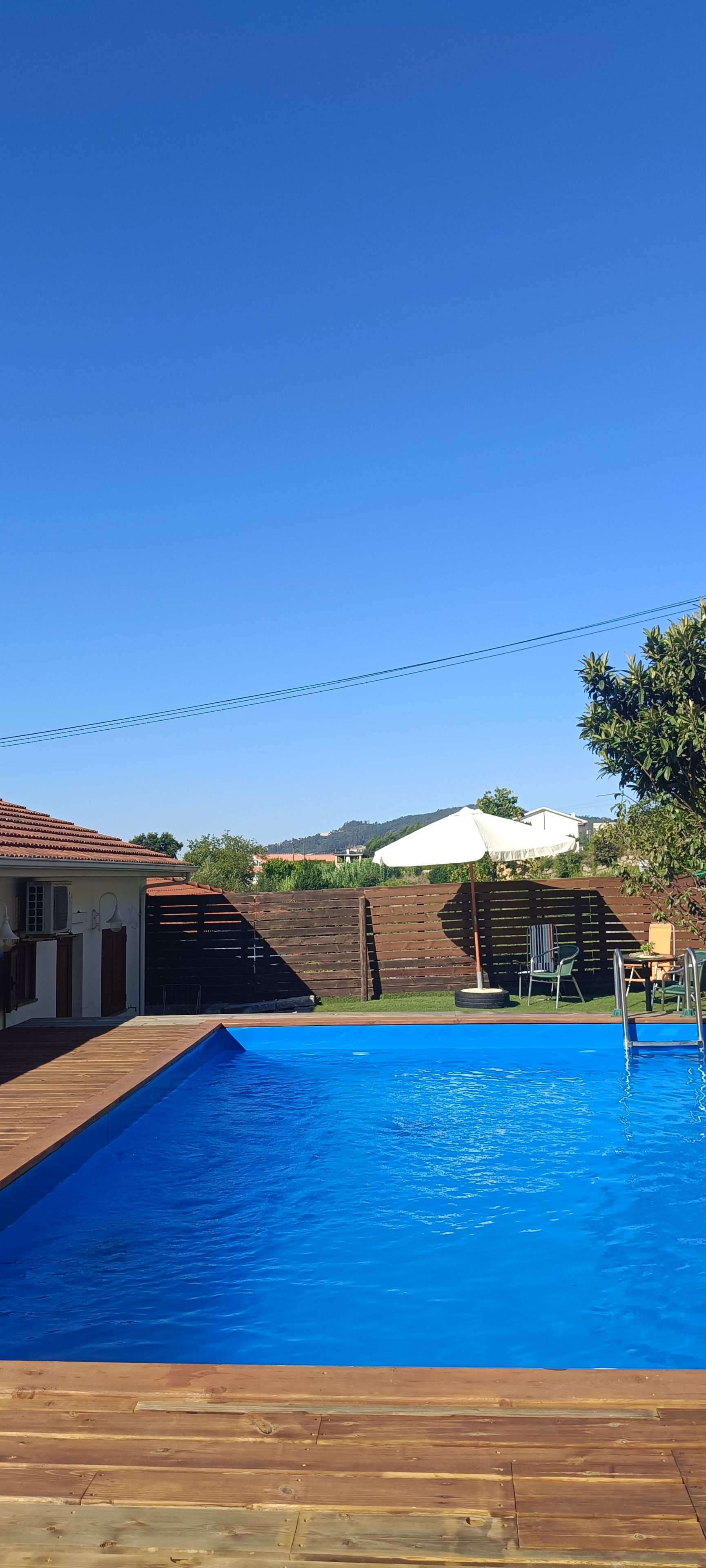 Ein Schwimmbecken mit Holzterrasse, einem weißen Sonnenschirm und Blick auf eine Hügelkuppe.