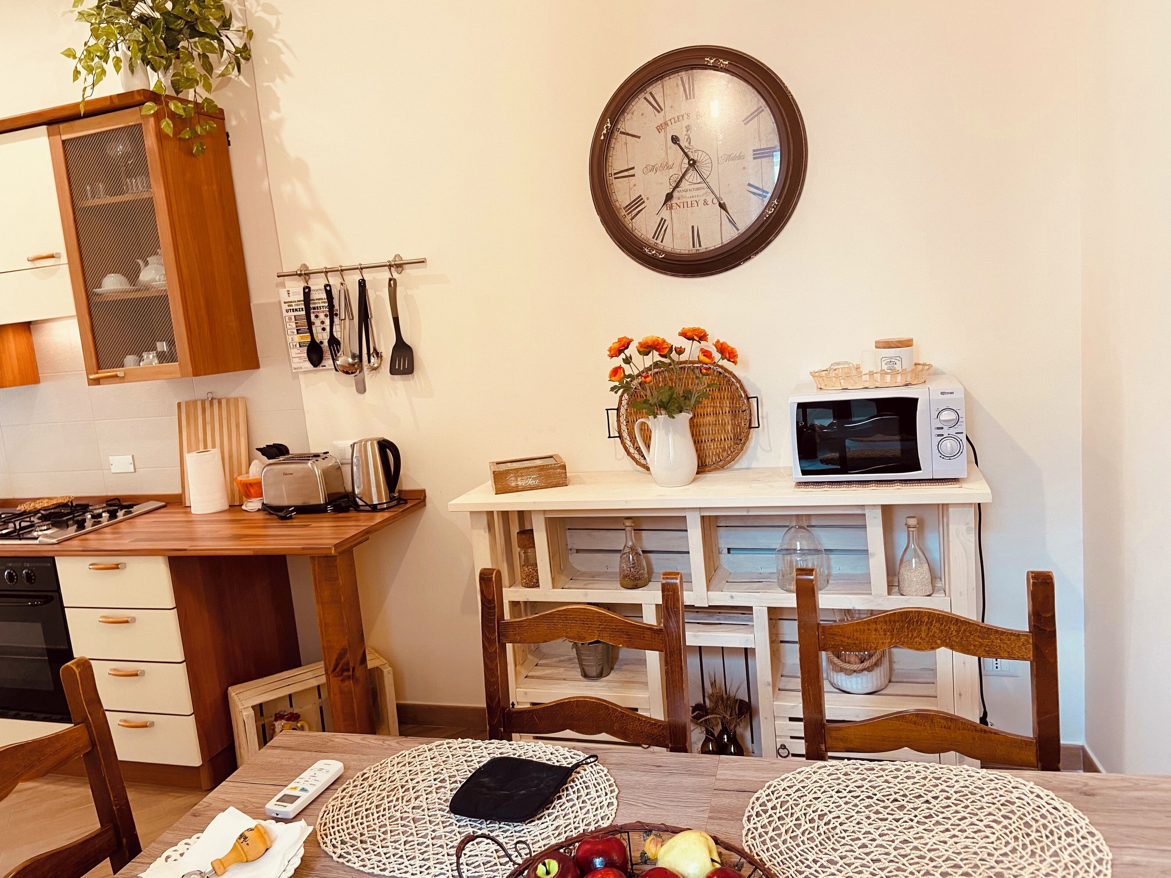 Una cocina con armarios de madera, un reloj, un microondas y una mesa de comedor con sillas.