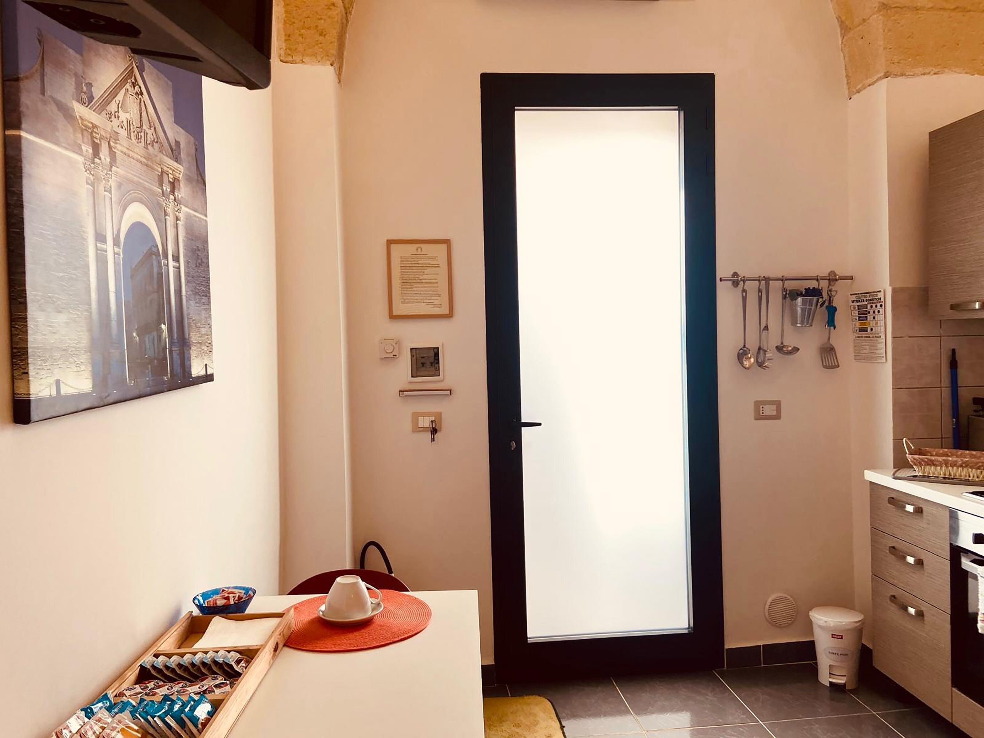 A narrow hallway with a door leading to a kitchen area. There is a framed picture on the wall, a small table with a cup and a tray of snacks, and a trash bin near the door.
