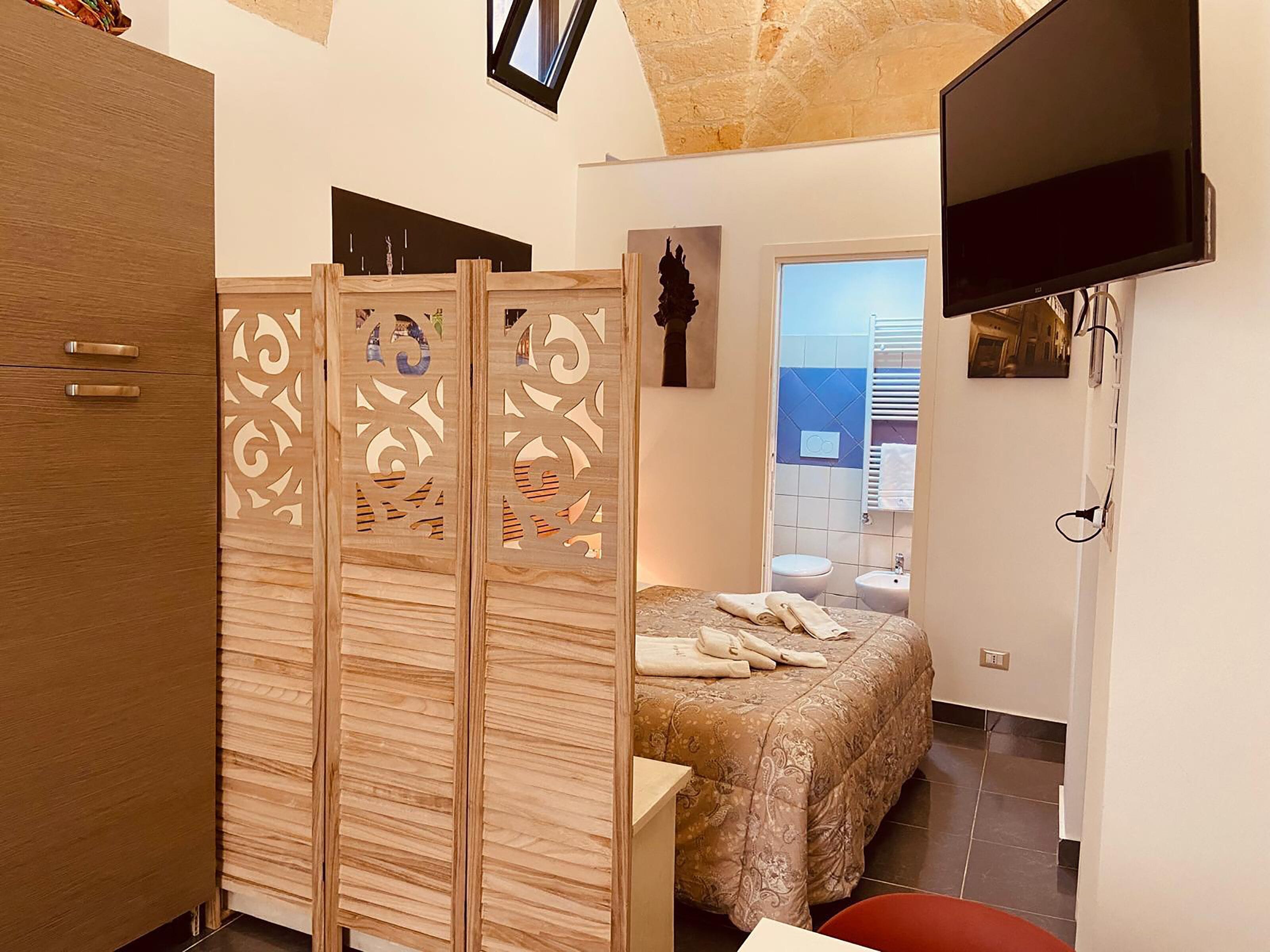 A bedroom with a bed, a TV, a wooden partition, and a bathroom visible through an open door.