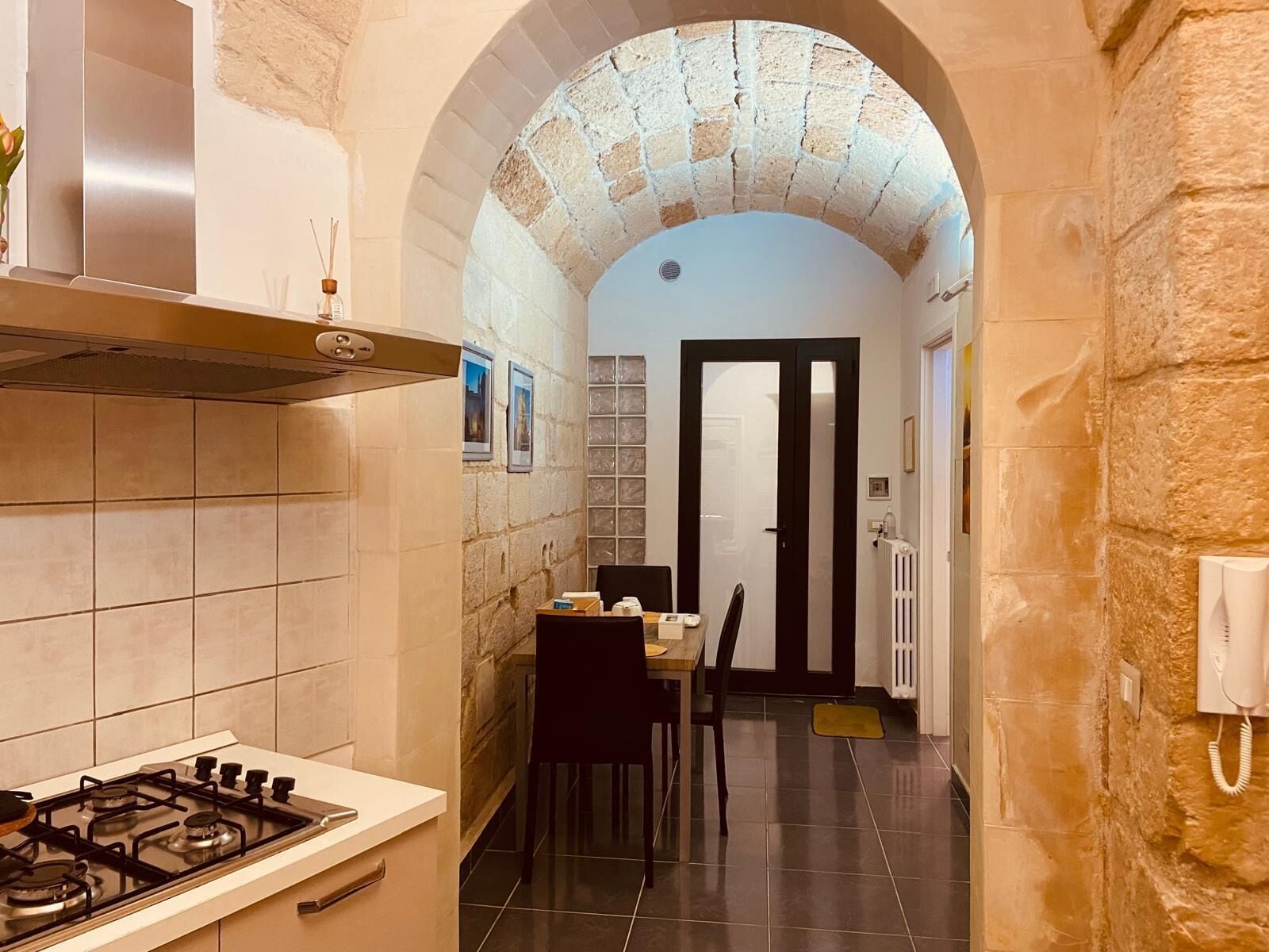 A narrow kitchen with a stone archway, a gas stove, a dining table with chairs, and a door leading to another room.
